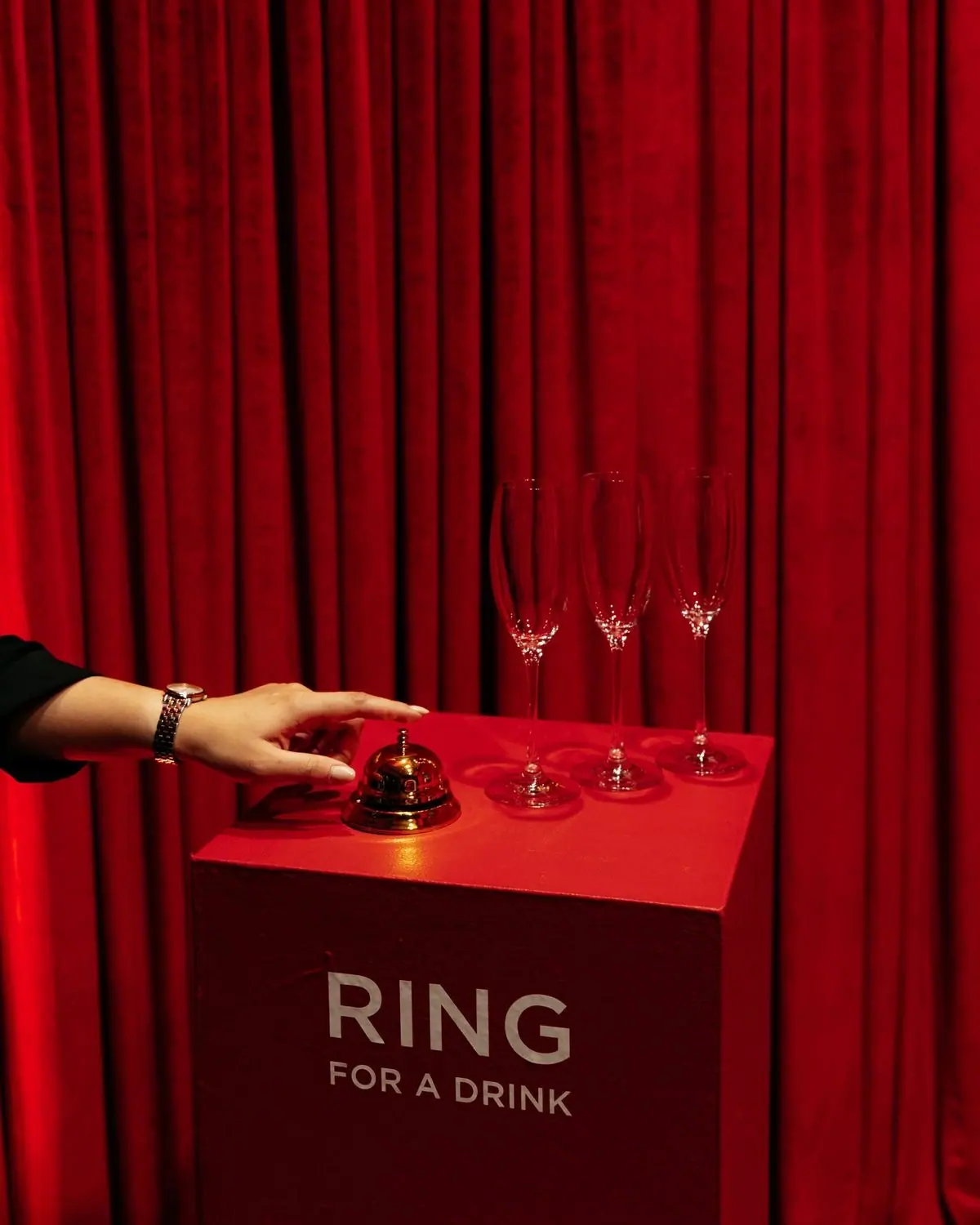 A hand rings a brass service bell beside three champagne glasses, against a red velvet curtain.