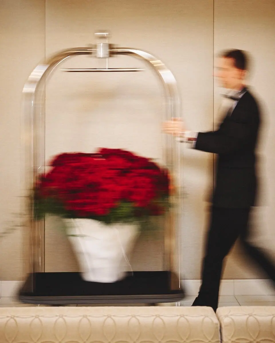 A bellman in a dark suit wheels a luggage cart of red flowers through a hotel lobby.