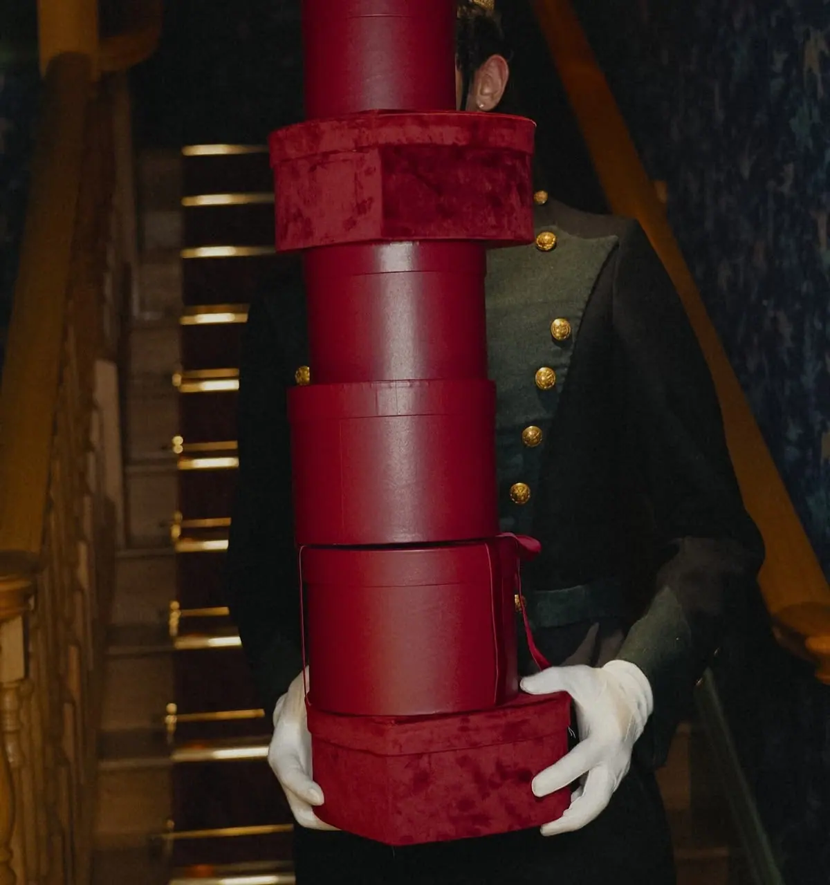 A bellboy in white gloves carries a tall stack of burgundy gift boxes down a staircase.