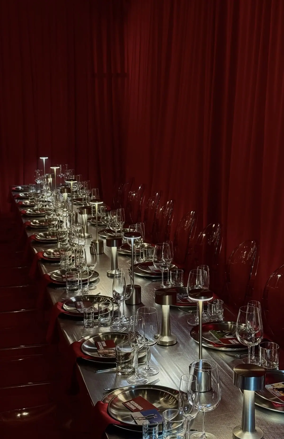 A long table set with glasses and small lamps along a wall of red curtains.