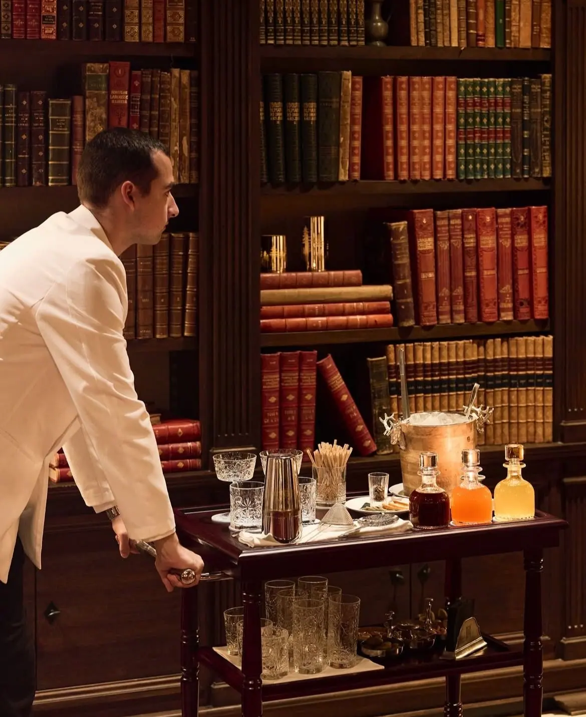 A host in a white jacket brings a drinks trolley through a private library.