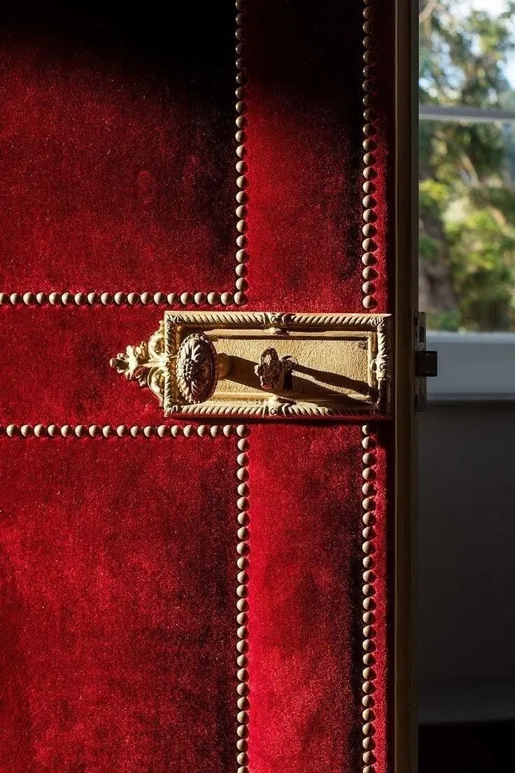 A red velvet door with brass studs and an ornate handle, beside a window onto a garden.